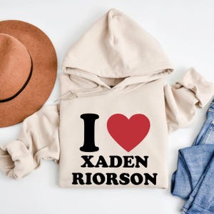 May include: A beige hoodie with the text "I ❤️ XADEN RIORSON" in black and red. The hoodie has a drawstring hood and long sleeves. A brown hat and blue jeans are partially visible in the background.
