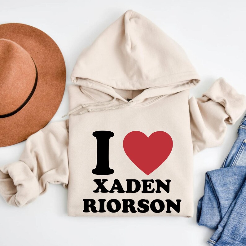 May include: A beige hoodie with the text "I ❤️ XADEN RIORSON" in black and red. The hoodie has a drawstring hood and long sleeves. A brown hat and blue jeans are partially visible in the background.