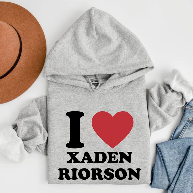 May include: A light gray hooded sweatshirt with the text "I ❤️ XADEN RIORSON" in black and red. The sweatshirt has a drawstring hood and long sleeves. The image also shows a brown hat and blue jeans.