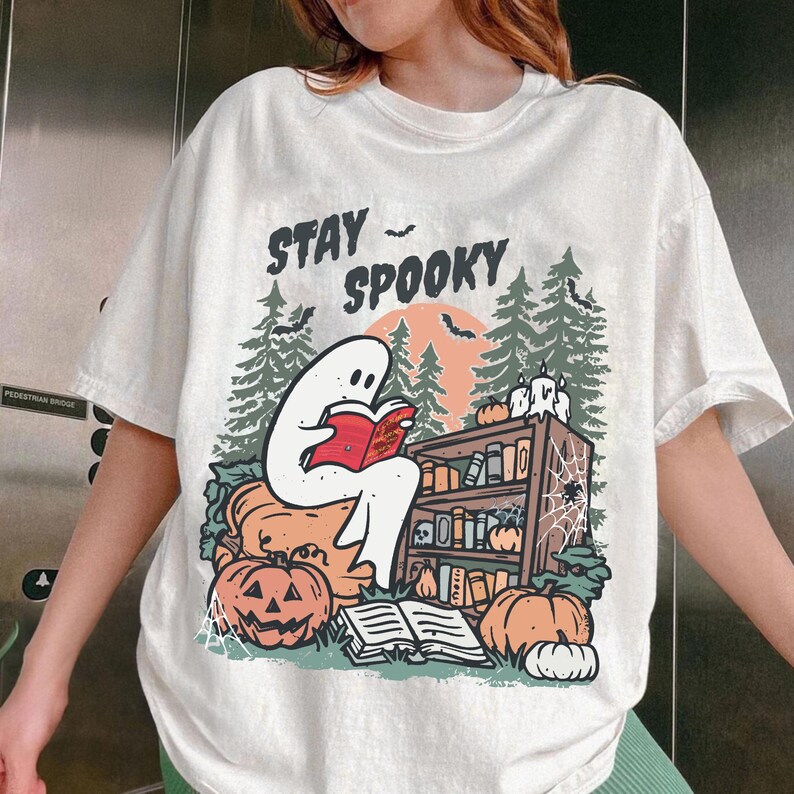 May include: White t-shirt with a Halloween graphic. The design shows a ghost reading a book, pumpkins, a bookshelf, and the words "Stay Spooky". The illustration uses orange, green, and black colours.