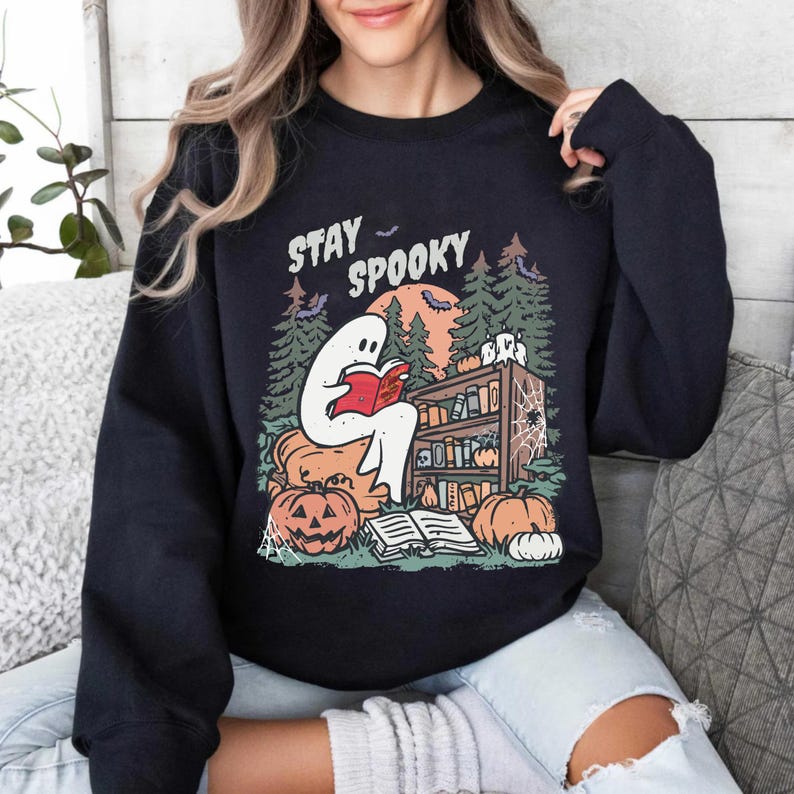 May include: Black sweatshirt with a Halloween graphic. The design shows a ghost reading a book, a bookshelf, pumpkins, and a full moon. The words "Stay Spooky" are printed above the ghost.