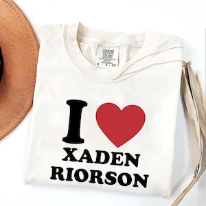 May include: Cream-colored t-shirt with the text "I ❤️ XADEN RIORSON" in black and red. The shirt features a black letter "I" followed by a red heart symbol. The text "XADEN RIORSON" is printed in a bold, sans-serif font.