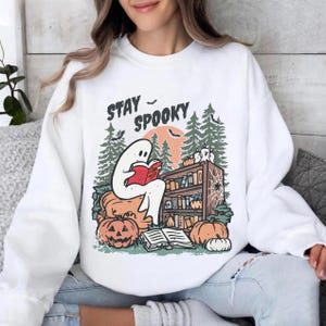 May include: White sweatshirt with a Halloween graphic. The design shows a ghost reading a book, a bookshelf, pumpkins, and the text "Stay Spooky". The background includes trees, a moon, and bats.