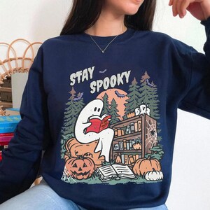 May include: Navy blue sweatshirt with a Halloween graphic. The design shows a white ghost reading a book in front of a bookshelf, pumpkins, and the text "Stay Spooky". Bats and trees complete the spooky scene.