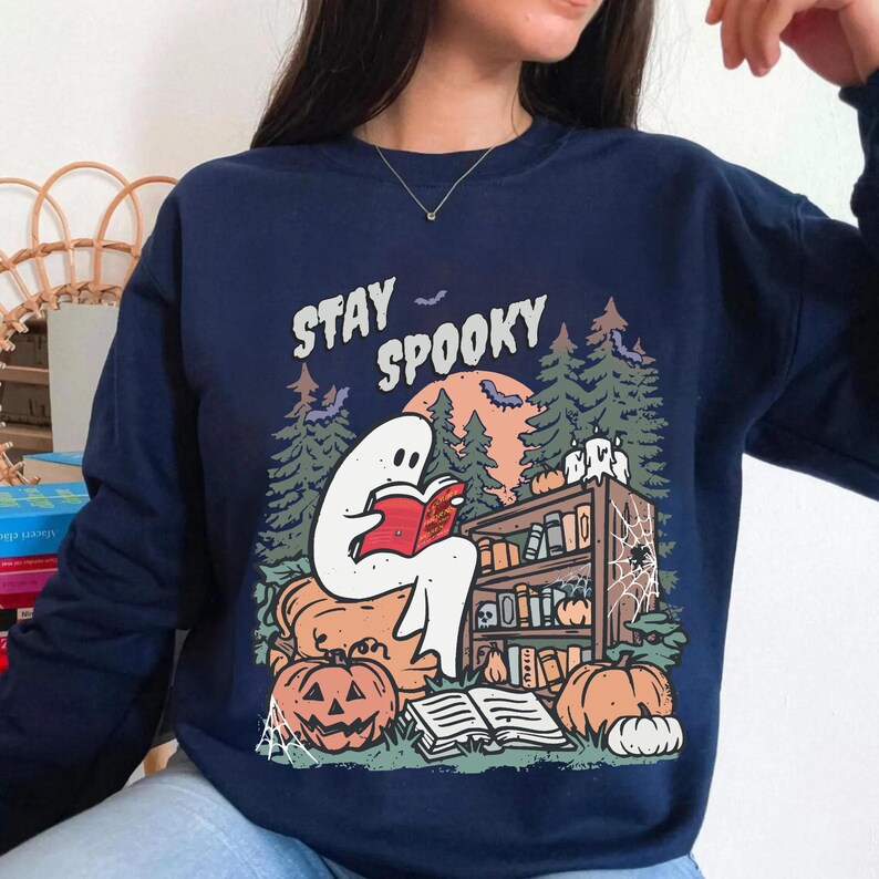 May include: Navy blue sweatshirt with a Halloween graphic. The design shows a white ghost reading a book in front of a bookshelf, pumpkins, and the text "Stay Spooky". Bats and trees complete the spooky scene.
