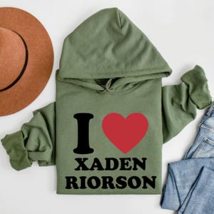 May include: An olive green hoodie with the text "I" and a red heart, followed by "XADEN RIORSON" in black. The hoodie has a hood and long sleeves. A brown hat and blue jeans are partially visible in the background.