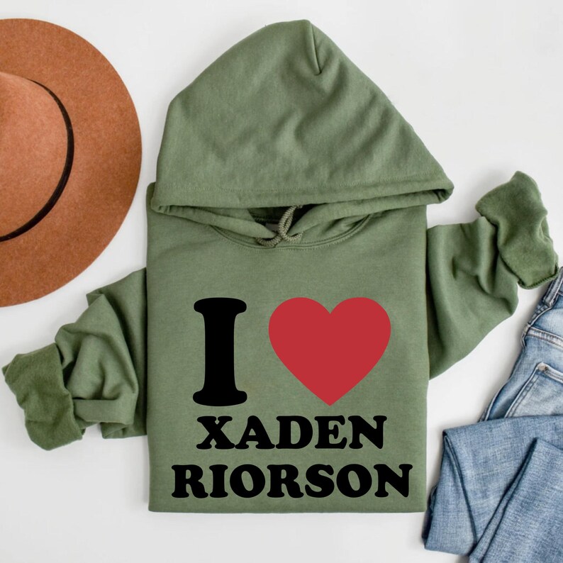 May include: An olive green hoodie with the text "I" and a red heart, followed by "XADEN RIORSON" in black. The hoodie has a hood and long sleeves. A brown hat and blue jeans are partially visible in the background.
