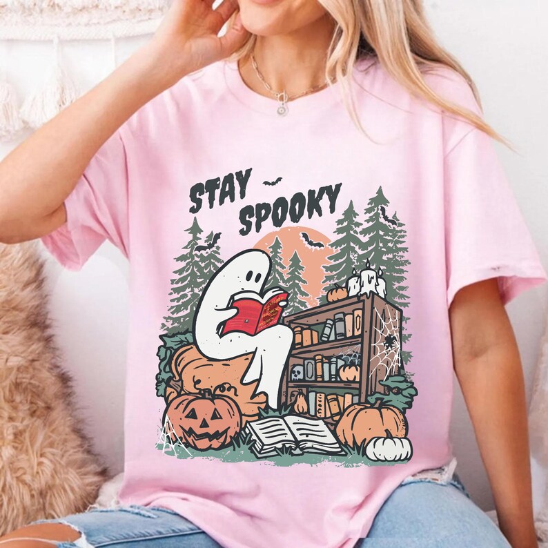 May include: Pale pink t-shirt with a Halloween graphic. The design shows a ghost reading a book, with pumpkins, a bookshelf, and the words "Stay Spooky". The background includes trees and a full moon.