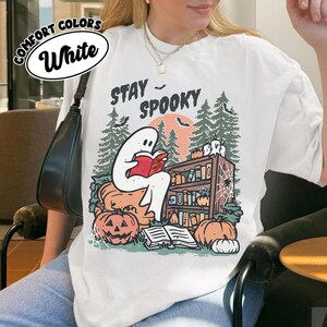 May include: White Comfort Colors t-shirt with a Halloween graphic. The design shows a ghost reading a book, pumpkins, a bookshelf, and the words "Stay Spooky".