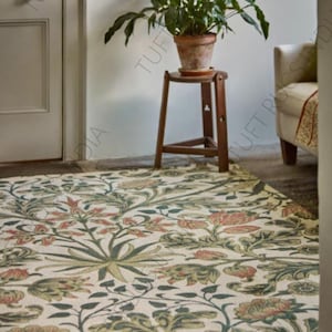 May include: A floral patterned rug with a cream background and a design of green leaves and red flowers. A small wooden stool holds a potted plant. The rug is in a room with a white door and a cream-colored chair.