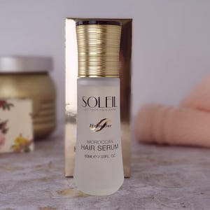 May include: A frosted glass bottle of Soleil Moroccan Hair Serum with a gold cap and a gold box in the background. The bottle has the text "SOLEIL LET YOUR HAIR SHINE" and "60ml 2.03 fl oz".