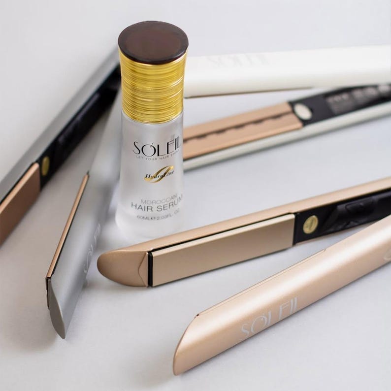 May include: A bottle of Soleil Moroccan Hair Serum and several gold and black hair straighteners are arranged on a white surface. The serum bottle has a gold cap and a label with the brand name and product information. The straighteners have gold plates and black handles.