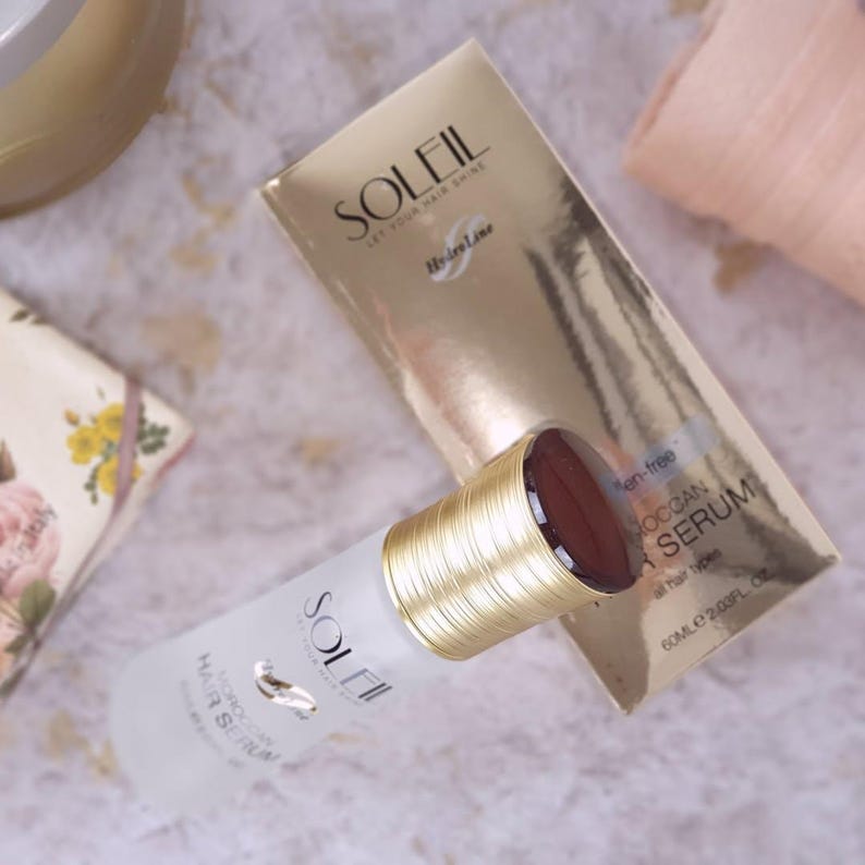 May include: A bottle of Soleil Moroccan Hair Serum with a gold cap and a matching gold box. The bottle is white with gold lettering and a gold emblem. The box is gold with the Soleil logo and product information. The serum is for all hair types.
