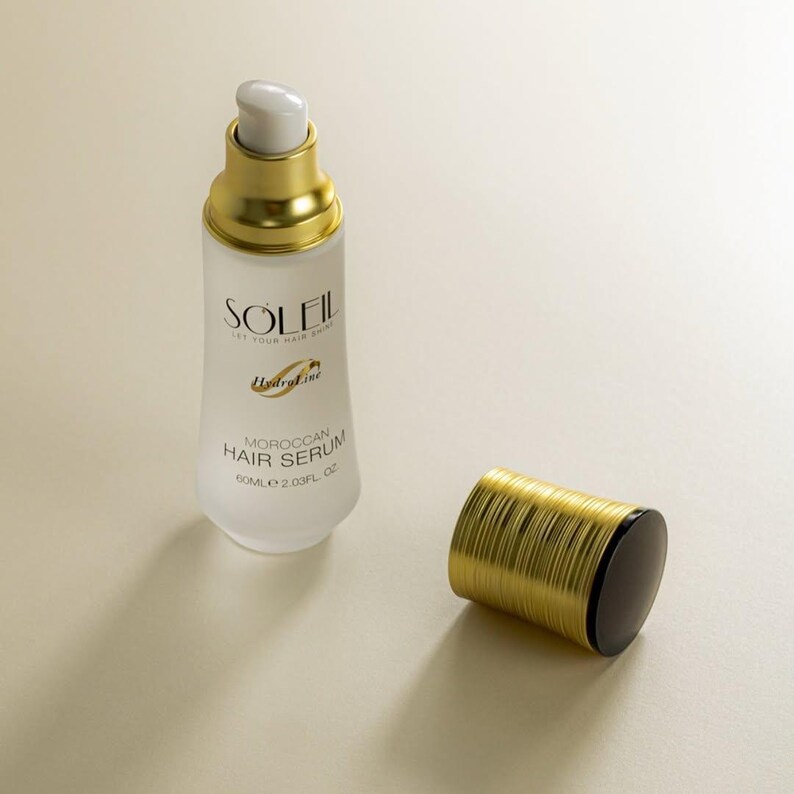 May include: A white and gold Moroccan Hair Serum bottle with the text "SOLEIL" and "Moroccan Hair Serum". The bottle is next to its gold and black cap. The bottle contains 2.03 fl oz of serum.