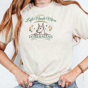 Può includere: T-shirt color crema con grafica di un Dobermann e la scritta "Life Needs More Dobermans". Il design include dettagli verdi e oro. La maglietta è realizzata in un materiale morbido e confortevole.