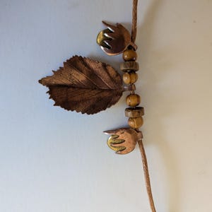 Puede incluir: Colgante de cobre artesanal con una hoja detallada y dos capullos florales estilizados. Ensartado en un cordón marrón, con cuentas de madera. El cobre tiene un acabado cálido y natural, ideal para joyería de inspiración natural.