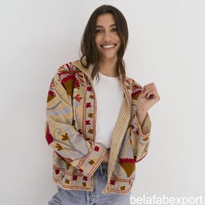 Pode incluir: Casaco bege com bordados florais em vermelho, castanho e azul. O casaco tem forro listrado e capuz. As mangas são compridas e o casaco é aberto na frente. O texto "belafabexport" é visível no canto inferior direito.