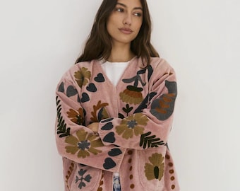 Puede incluir: Chaqueta rosa claro con frente abierto y bordados florales en tonos marrón, verde y gris. La chaqueta tiene dos bolsillos y está hecha de una tela suave y texturizada. El diseño floral incluye formas de corazón y es visible en las mangas y el frente.