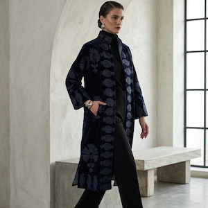 Könnte beinhalten: Ein langer, marineblauer Samtmantel mit Stehkragen und aufwendiger, hellblauer Blumen- und Granatapfelstickerei entlang der vorderen Knopfleiste, der Ärmel und des Saums. Der Mantel wird über einem schwarzen Rollkragenpullover und einer Hose getragen.