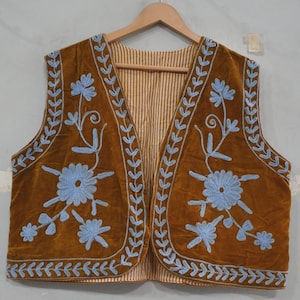 May include: A brown velvet waistcoat with light blue floral embroidery and leaf-like trim. The waistcoat has a striped lining and is displayed on a wooden hanger. The embroidery features stylized flowers and vines.