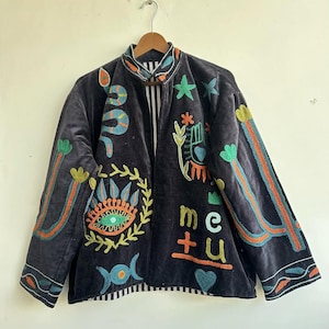 May include: Dark gray velvet jacket with colorful embroidered symbols, including a snake, eye, hand, and the letters "me + u." The jacket has a mandarin collar and striped lining, with orange, green, and blue accents.