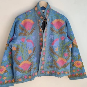 May include: A light blue jacket with intricate floral embroidery in pink, orange, and blue. The jacket features a high collar and long sleeves, with a detailed pattern of flowers and leaves. The jacket is hanging on a wooden hanger.