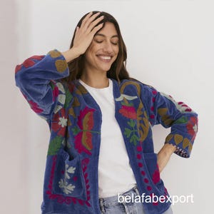 Puede incluir: Chaqueta de terciopelo azul con bordados florales coloridos. La chaqueta tiene un corte relajado, dos bolsillos delanteros y mangas tres cuartos. El bordado presenta detalles en rosa, rojo, verde y dorado. El texto "belafabexport" es visible en la parte inferior derecha.