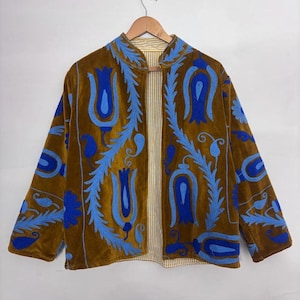 May include: A brown velvet jacket with blue floral embroidery. The jacket has a collarless design and long sleeves. The interior lining is striped. The jacket is hanging on a wooden hanger.