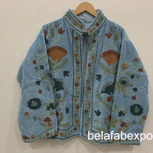 May include: A light blue jacket with intricate floral embroidery in shades of orange, green, and brown. The jacket has a high collar and is displayed on a wooden hanger. The text "belafabexport" is visible at the bottom right.