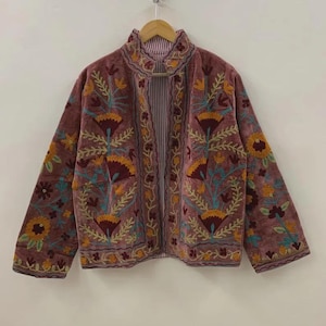 May include: A dusty rose jacket with intricate floral embroidery in shades of yellow, orange, teal, and burgundy. The jacket has a high collar and is lined with a striped fabric. The sleeves are long and the jacket is hanging on a wooden hanger.