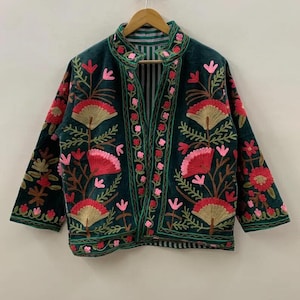 Può includere: Giacca ricamata verde scuro con collo alla coreana e maniche a tre quarti. La giacca presenta un motivo floreale nei toni del rosso, rosa e oro. La giacca è appesa a una gruccia di legno.