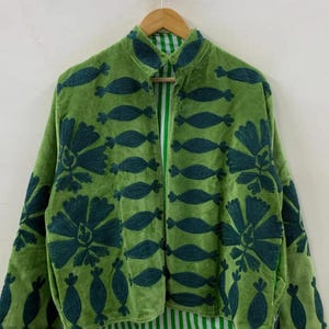 May include: A green velvet jacket with dark teal floral and abstract embroidered designs. The jacket has a high collar and a striped green and white lining. The jacket is hanging on a wooden hanger.