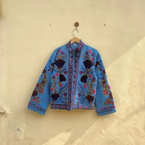 May include: A light blue jacket with intricate floral embroidery in red, brown, and green. The jacket features a mandarin collar and long sleeves. The embroidery creates a striking contrast against the blue fabric. The jacket is hanging on a wooden hanger.
