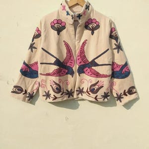 May include: Cream-coloured jacket featuring embroidered pink and blue swallows and floral designs. The jacket has a high collar and a full-length zip. The embroidery includes pink flowers and blue accents, creating a folk art style.