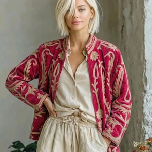 Puede incluir: Una chaqueta de terciopelo magenta vibrante con intrincados bordados en tono dorado en un patrón floral en espiral, combinada con una camisa abotonada de color beige claro y pantalones con cordón a juego, creando un conjunto de manga larga.