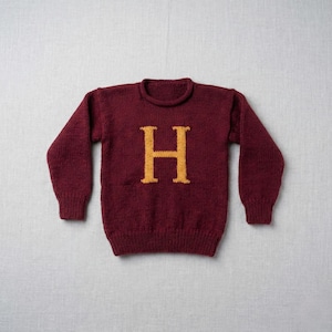 Puede incluir: Un suéter de punto burdeos con una gran letra "H" dorada en la parte delantera. El suéter tiene mangas largas y cuello redondo. La prenda de punto es de color liso, con dobladillo y puños acanalados.