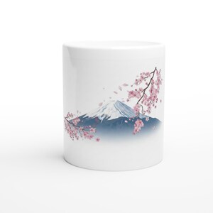 以下が含まれることがあります： 白いセラミックマグカップ。水彩画で描かれた富士山のイラストが特徴で、雪をかぶった山頂と青灰色のベースが描かれています。ピンク色の桜の枝と舞い散る花びらが山を彩っています。