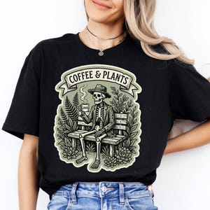 Può includere: T-shirt nero con grafica di uno scheletro con cappello, seduto su una panchina, che tiene una tazza di caffè, circondato da piante. Il testo "COFFEE & PLANTS" è sopra l'immagine in uno striscione. La maglietta è abbinata a jeans blu.