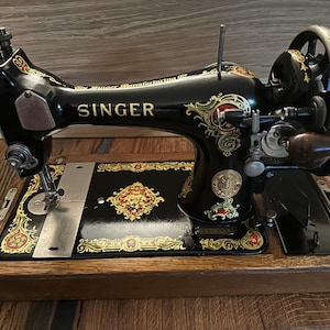 May include: An antique Singer sewing machine, black with gold and red decorative accents. The machine features the Singer logo and ornate floral designs. It is mounted on a wooden base.
