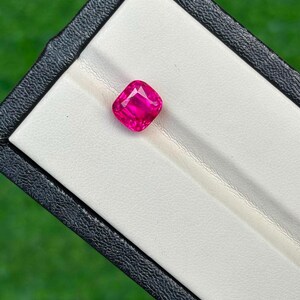 May include: A vibrant, cushion-cut pink gemstone with a faceted surface that reflects light. The gemstone is securely positioned within a white velvet-lined slot inside a black rectangular display box, showcasing its intense color and clarity.