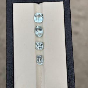 May include: Four faceted, light blue, translucent gemstones arranged vertically in a rectangular display box. The box features a dark brown outer frame and a cream-coloured interior insert with a central groove to hold the stones securely in place.