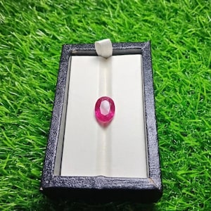 May include: A faceted oval-shaped pink gemstone centered on a white velvet insert within a rectangular black display box. The gemstone exhibits a vibrant pink hue with clear light reflections across its polished surface, presented in a secure case.