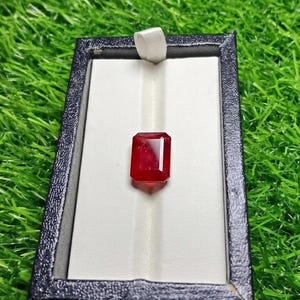 May include: A vibrant red gemstone with an emerald cut, centered within a rectangular black display box featuring a white interior lining and a small white fabric pull tab for opening the lid, presented as a single loose stone for display.