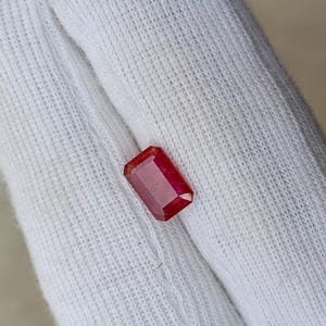 May include: A rectangular, faceted ruby gemstone with a deep red hue. The gem is set against a white fabric background, showcasing its clarity and color. The cut of the stone highlights its brilliance.
