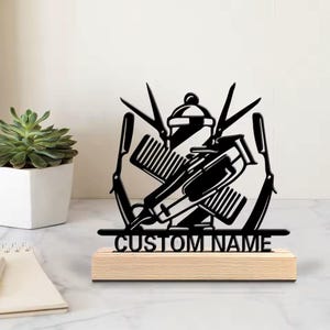 Könnte beinhalten: Schwarzes Metall-Friseurschild mit gekreuzten Friseurwerkzeugen, darunter Schere, Rasierer und Haarschneider. Das Schild ist auf einem hellen Holzsockel mit dem Text "CUSTOM NAME" montiert. Ein dekorativer Artikel für einen Friseursalon.