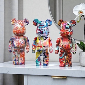 Peut inclure: Trois figurines en forme d'ours avec des motifs de style graffiti colorés. Les figurines sont exposées sur une surface blanche. Chaque ours présente un motif unique de couleurs vives, notamment le rouge, le bleu, le jaune et le blanc. Les ours ont des oreilles arrondies et un corps carré.