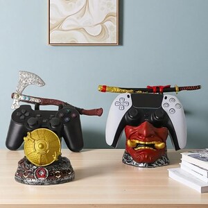 Könnte beinhalten: Zwei Gaming-Controller-Ständer. Ein Ständer hat einen schwarzen Controller mit einem goldenen Schild und einer Axt. Der andere Ständer hat einen weiß-schwarzen Controller mit einer roten Dämonenmaske und einem Katana-Schwert.