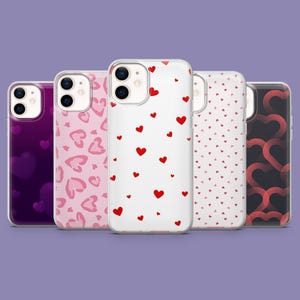 Può includere: Una collezione di cinque custodie per telefoni con vari disegni a forma di cuore. Le custodie presentano motivi di cuori rossi su bianco, cuori rosa con stampa leopardata e motivi a cuore sfumati in rosa e viola. Su sfondo viola.