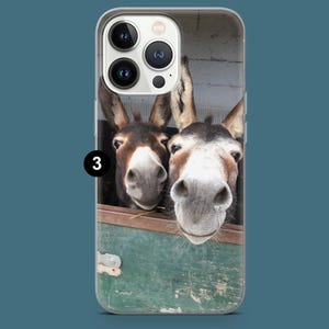 May include: A phone case displaying a photograph of two donkeys. They are looking over a green wooden barrier. The donkeys have brown and white fur, with their noses close to the camera. The case has a clear border and a blue background.
