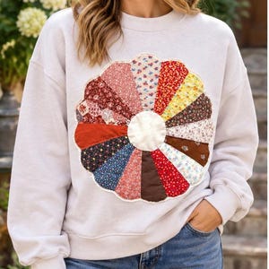 May include: A light beige crewneck sweatshirt featuring a large, circular, scalloped-edge applique on the chest. The applique is constructed from multiple wedge-shaped fabric segments in various floral and solid patterns of red, blue, and yellow.
